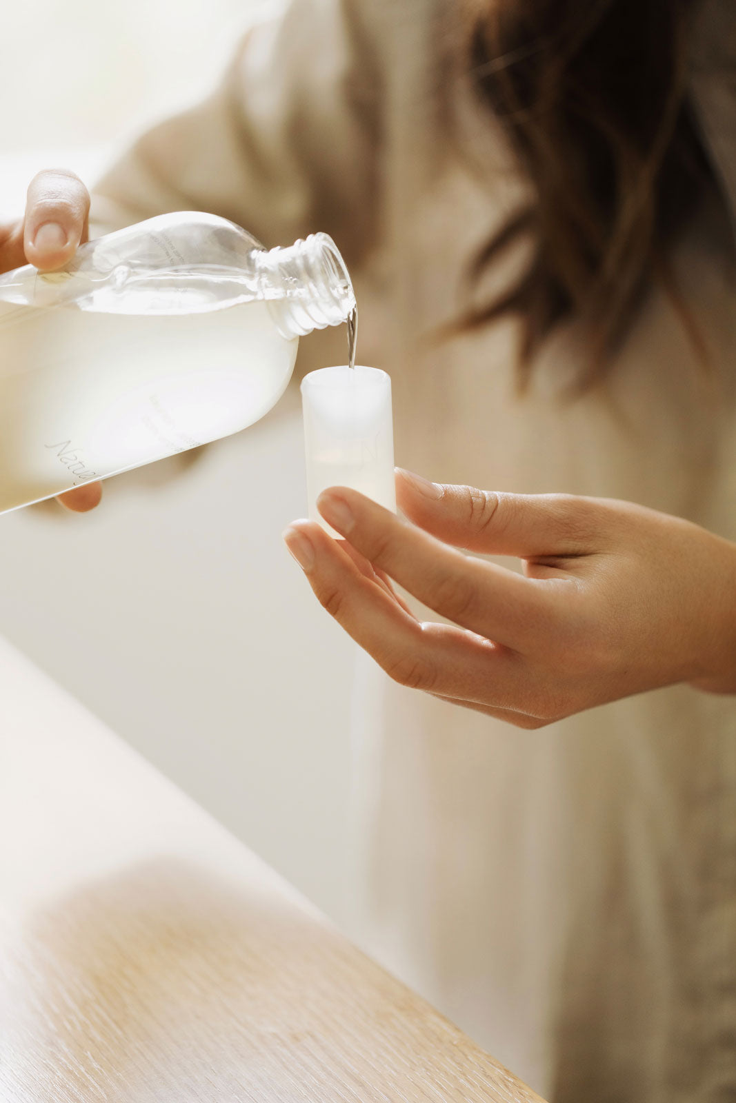 Person pouring a liquid from a bottle into a container on a light wooden surface.
