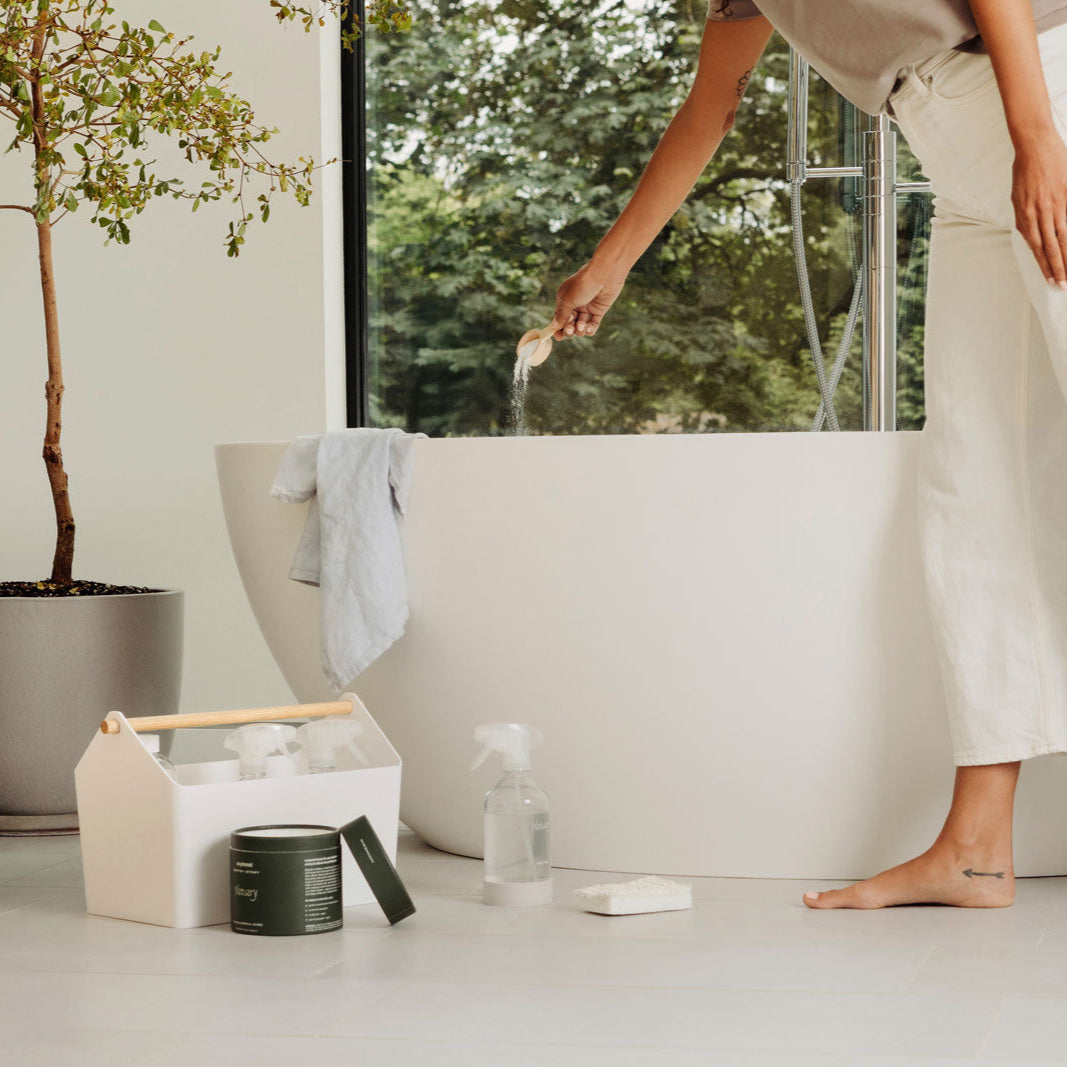 Woman standing by a white bathtub with various natuary products on a light-colored floor.